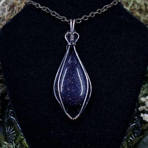 May include: A teardrop-shaped pendant necklace with a dark blue, glittery stone encased in a silver-colored wire frame. The pendant hangs from a delicate chain. The necklace is displayed on a black bust against a backdrop of greenery.