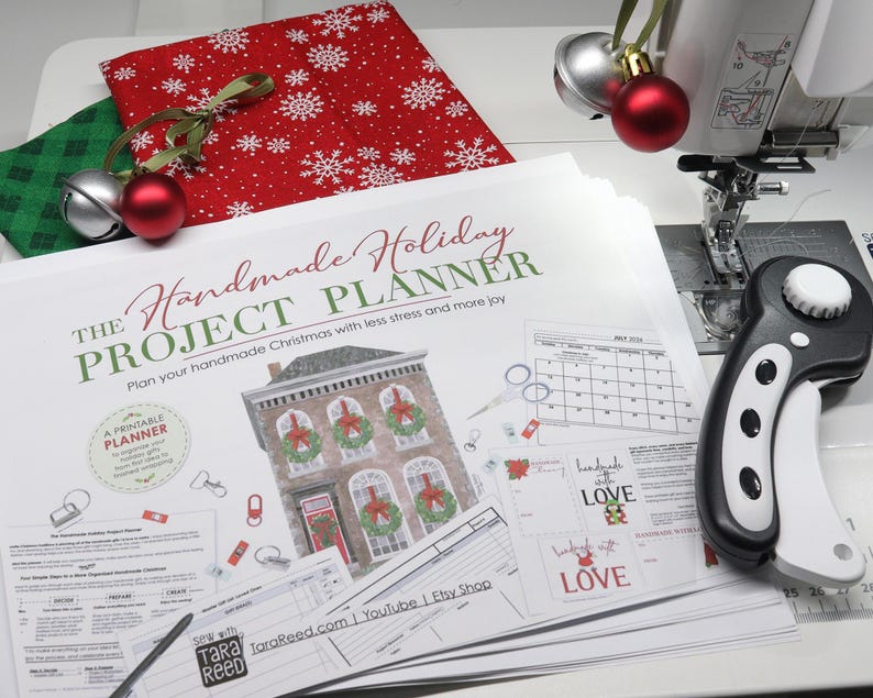 Printed Handmade Holiday Project Planner beside a sewing machine, rotary cutter, Christmas fabric, and jingle bells, ready for planning handmade holiday gifts.