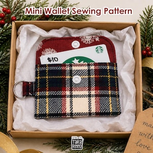 May include: A mini wallet sewing pattern featuring a red, navy blue, and cream plaid fabric exterior with a white snap closure and a metal D-ring. The wallet is displayed inside a cardboard box lined with white tissue paper, holding a gift card.