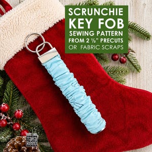 May include: A light blue fabric scrunchie key fob with a silver metal hardware clasp and a circular ring. The text on the image reads: SCRUNCHIE KEY FOB SEWING PATTERN FROM 2 1/2" PRECUTS OR FABRIC SCRAPS. The fabric features a subtle pattern.