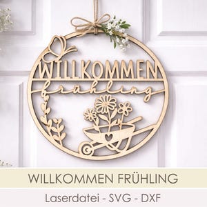 Könnte beinhalten: Ein kreisförmiges Holzschild mit den ausgeschnittenen Worten "WILLKOMMEN Frühling". Das Schild zeigt einen Schmetterling, Blumen in einer Schubkarre und Grünzeug. Es wird von einer Schnurschleife gehalten und mit kleinen weißen Blumen verziert.