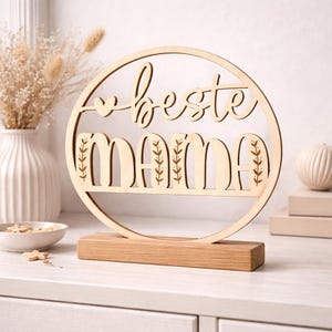 K&ouml;nnte beinhalten: Ein dekoratives Holzschild mit den Worten "beste mama" in einem kreisf&ouml;rmigen Design ausgeschnitten. Das Schild ist auf einem rechteckigen Holzsockel montiert. Die Buchstaben "M" und "A" haben blattartige Designs. Das Schild ist hellbraun.