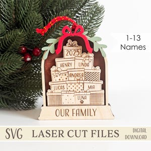 Puede incluir: Adorno navideño de madera con una pila de cajas de regalo con nombres y el año 2025. El adorno tiene un lazo rojo y una cuerda roja y verde para colgar. El texto "OUR FAMILY" está en la parte inferior.