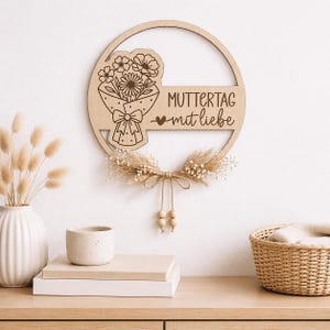 May include: A wooden Mother's Day sign with a bouquet of flowers and the words "MUTTERTAG mit liebe" engraved. The sign is circular with a dried floral arrangement and wooden beads. The sign is hanging on a white wall.