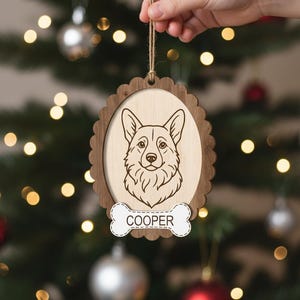 May include: Wooden Christmas ornament featuring a detailed engraved corgi portrait within an oval frame. The frame has a scalloped edge and a bone-shaped tag with the name "COOPER". The ornament hangs from a twine string, perfect for pet lovers.