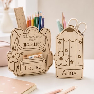 Puede incluir: Dos organizadores de escritorio de madera clara con forma de mochila y l&aacute;piz. La mochila presenta el texto Alles Gute zur EINSCHULUNG y Louise, mientras que el l&aacute;piz presenta el nombre Anna. Ambos art&iacute;culos est&aacute;n decorados con motivos florales.