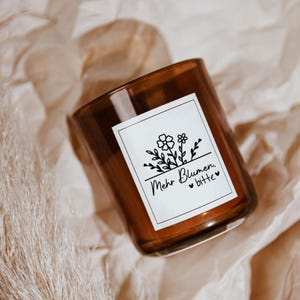 Pode incluir: Um frasco de vela de vidro castanho com um r&oacute;tulo branco com um desenho floral e o texto "Mehr Blumen, bitte" em preto. O frasco da vela est&aacute; sobre um fundo neutro e texturizado.