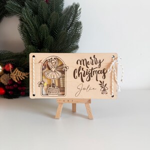 May include: Wooden Christmas sign with a ballerina illustration and the words "Merry Christmas Julie". The sign is displayed on a small wooden easel, with a green wreath in the background.