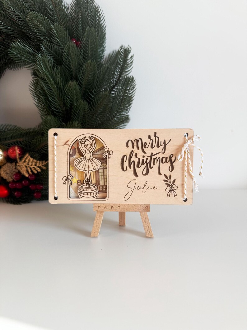 May include: Wooden Christmas sign with a ballerina illustration and the words "Merry Christmas Julie". The sign is displayed on a small wooden easel, with a green wreath in the background.