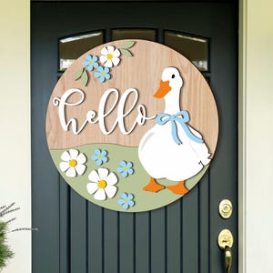 May include: A round wooden door sign with a white goose wearing a blue bow. The sign has the word "hello" in cursive, with white and blue flowers. The background is a light brown wood grain.