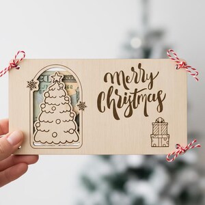 May include: A wooden Christmas gift card holder with a laser-cut Christmas tree design and the words "Merry Christmas." The holder is decorated with red and white twine and contains money. A Christmas tree is in the background.