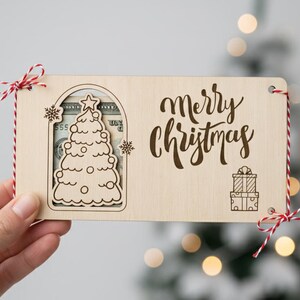 May include: A wooden Christmas gift card holder with a laser-cut Christmas tree design and the words "Merry Christmas". The holder is tied with red and white twine and a bow. A gift card is visible inside.