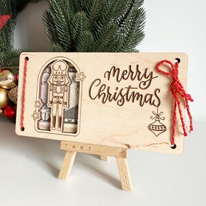 May include: A wooden Christmas card with a laser-cut nutcracker design and the words "Merry Christmas." The card is displayed on a small wooden easel. Red string is attached to the card.