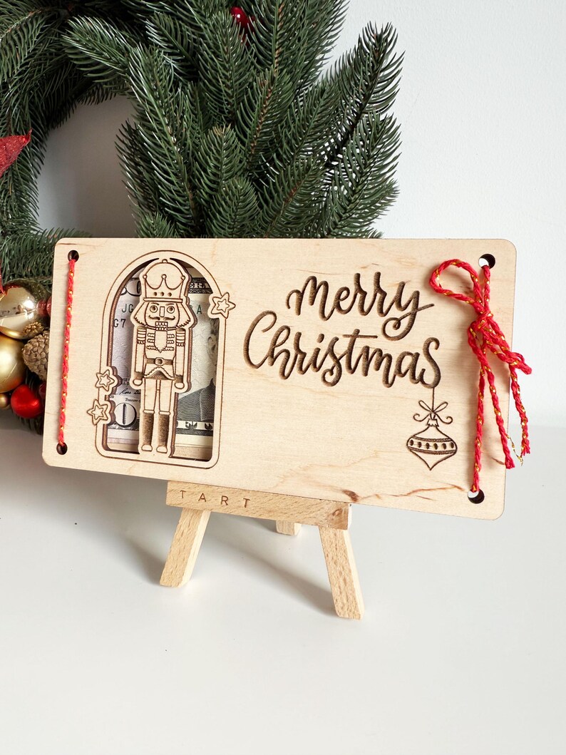 May include: A wooden Christmas card with a laser-cut nutcracker design and the words "Merry Christmas." The card is displayed on a small wooden easel. Red string is attached to the card.