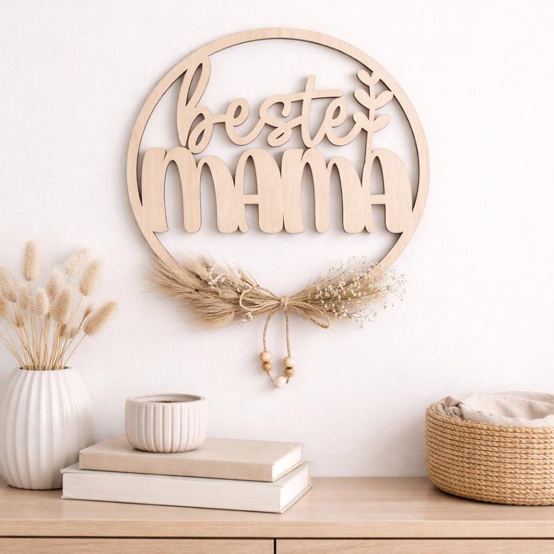K&ouml;nnte beinhalten: Ein h&ouml;lzernes Wandbild mit den Worten "Beste Mama" in Schreib- und Gro&szlig;buchstaben, umgeben von einem kreisf&ouml;rmigen Rahmen. Das Design umfasst Trockenblumen und eine Schnur mit Holzperlen. Es h&auml;ngt an einer wei&szlig;en Wand.
