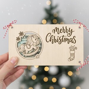 May include: A wooden Christmas gift card holder with a rocking horse design and the words "Merry Christmas". The holder is decorated with red and white twine and a Christmas tree is in the background.