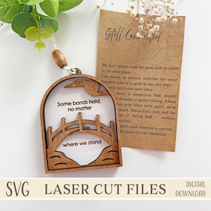 May include: A wooden laser-cut ornament with a bridge design and the words "Some bonds hold, no matter" and "where we stand." A small tag reads "Still Connected" with a poem. The ornament hangs from a white string.