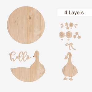 May include: Wooden wall decor set featuring a large round piece, a duck silhouette, the word "hello", and floral and bow accents. The pieces are made of light-colored wood and are designed to be layered.