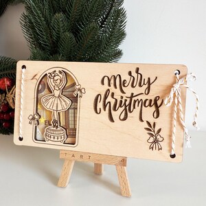 May include: Wooden Christmas ornament with a ballerina design and the words "Merry Christmas" engraved. The ornament is rectangular, with a cutout of a ballerina on a stage. White string is attached for hanging.
