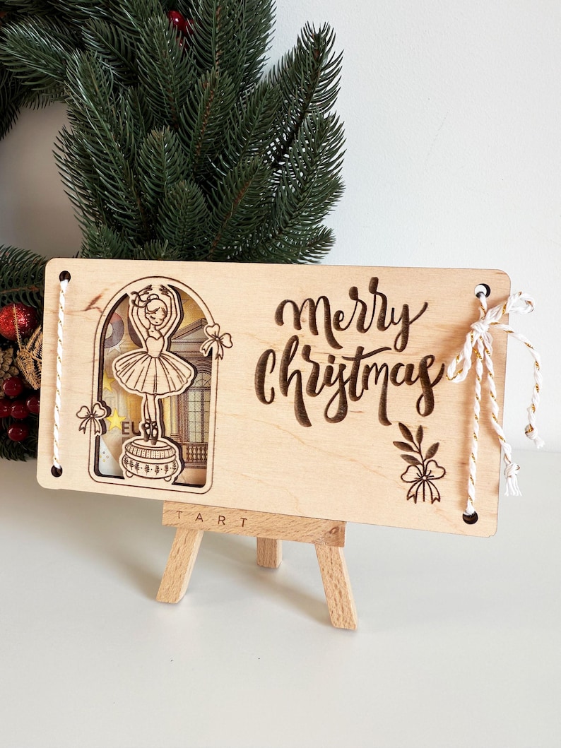 May include: Wooden Christmas ornament with a ballerina design and the words "Merry Christmas" engraved. The ornament is rectangular, with a cutout of a ballerina on a stage. White string is attached for hanging.