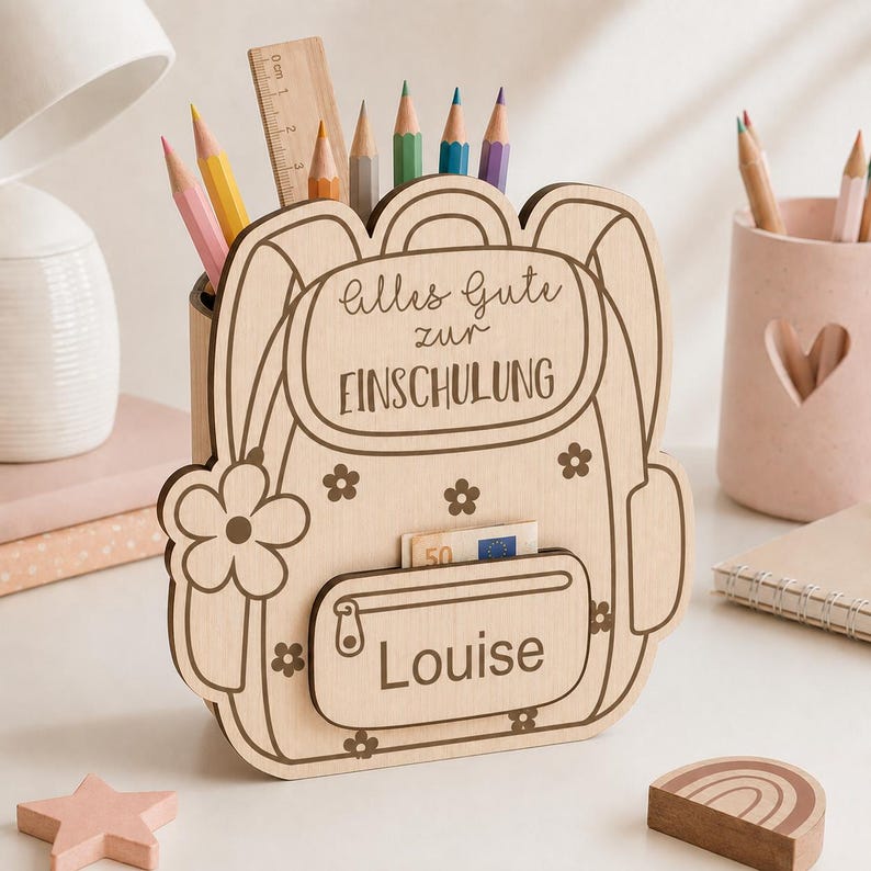 Puede incluir: Un portal&aacute;pices de madera clara con forma de mochila y dise&ntilde;o de flores. El texto "Alles Gute zur EINSCHULUNG" est&aacute; grabado en la solapa superior y el nombre "Louise" en el bolsillo delantero. Incluye una regla de madera en su interior.