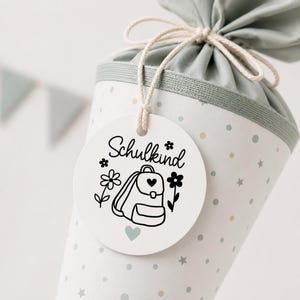 May include: A cone-shaped gift bag in light green with a pattern of small dots and stars, tied with a string. A round white tag hangs from the string, featuring the black text Schulkind, a backpack illustration, flowers, and a heart.