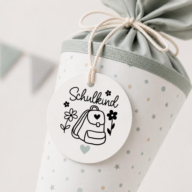 May include: A cone-shaped gift bag in light green with a pattern of small dots and stars, tied with a string. A round white tag hangs from the string, featuring the black text Schulkind, a backpack illustration, flowers, and a heart.