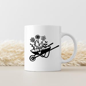 May include: White ceramic mug featuring a black illustration of a wheelbarrow filled with flowers and a heart. The wheelbarrow design is simple and elegant, perfect for gardening enthusiasts. The mug has a curved handle.