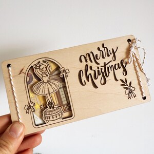 May include: Wooden Christmas card with a cutout design of a ballerina and the words "Merry Christmas". The card has a light wood finish and is decorated with string and bow accents.