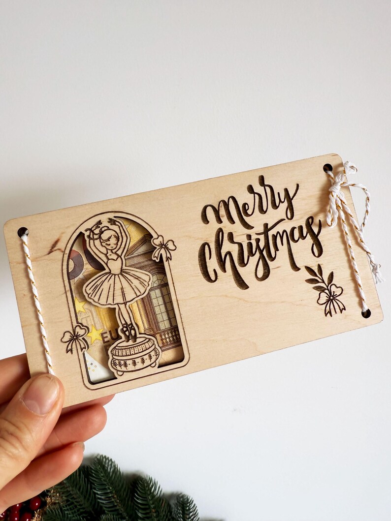 May include: Wooden Christmas card with a cutout design of a ballerina and the words "Merry Christmas". The card has a light wood finish and is decorated with string and bow accents.