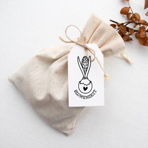 Pode incluir: Uma bolsa de linho bege amarrada com barbante, com uma etiqueta branca com um desenho a tra&ccedil;o preto de um bulbo de jacinto e a palavra "BLUMENZEIT". A bolsa est&aacute; sobre uma superf&iacute;cie branca com folhas secas.