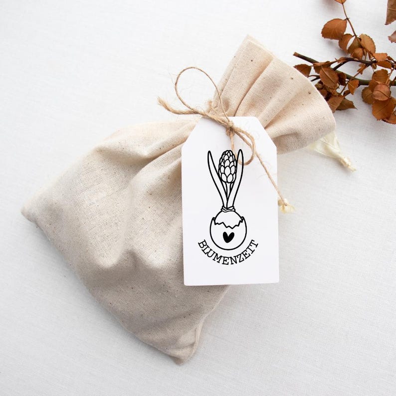 Pode incluir: Uma bolsa de linho bege amarrada com barbante, com uma etiqueta branca com um desenho a tra&ccedil;o preto de um bulbo de jacinto e a palavra "BLUMENZEIT". A bolsa est&aacute; sobre uma superf&iacute;cie branca com folhas secas.