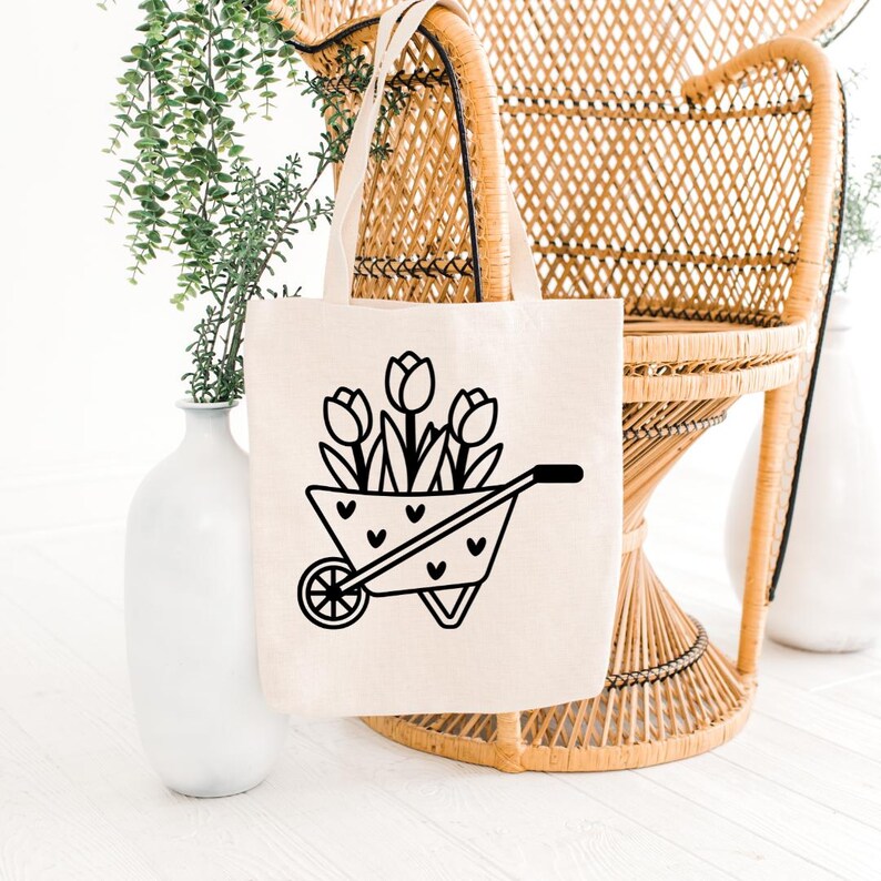 Pode incluir: Uma sacola bege com uma ilustra&ccedil;&atilde;o preta de tulipas em um carrinho de m&atilde;o com detalhes de cora&ccedil;&atilde;o. A sacola tem duas al&ccedil;as e &eacute; exibida em frente a uma cadeira de vime e vasos brancos com vegeta&ccedil;&atilde;o.