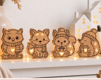 May include: Four light-up wooden animal figures including an owl on a branch, a fox, a squirrel holding an acorn, and a hedgehog holding a cup. Each figure features cut-out heart and leaf shapes that glow with warm light. Text reads: DIGITALE DATEI - SVG - DXF.