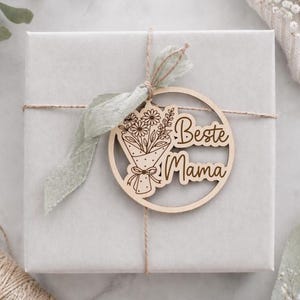 May include: A light gray gift box tied with twine and a sage green ribbon. A wooden circular tag with a laser-cut bouquet of flowers and the words "Beste Mama" is attached to the box. The tag is made of light-colored wood.