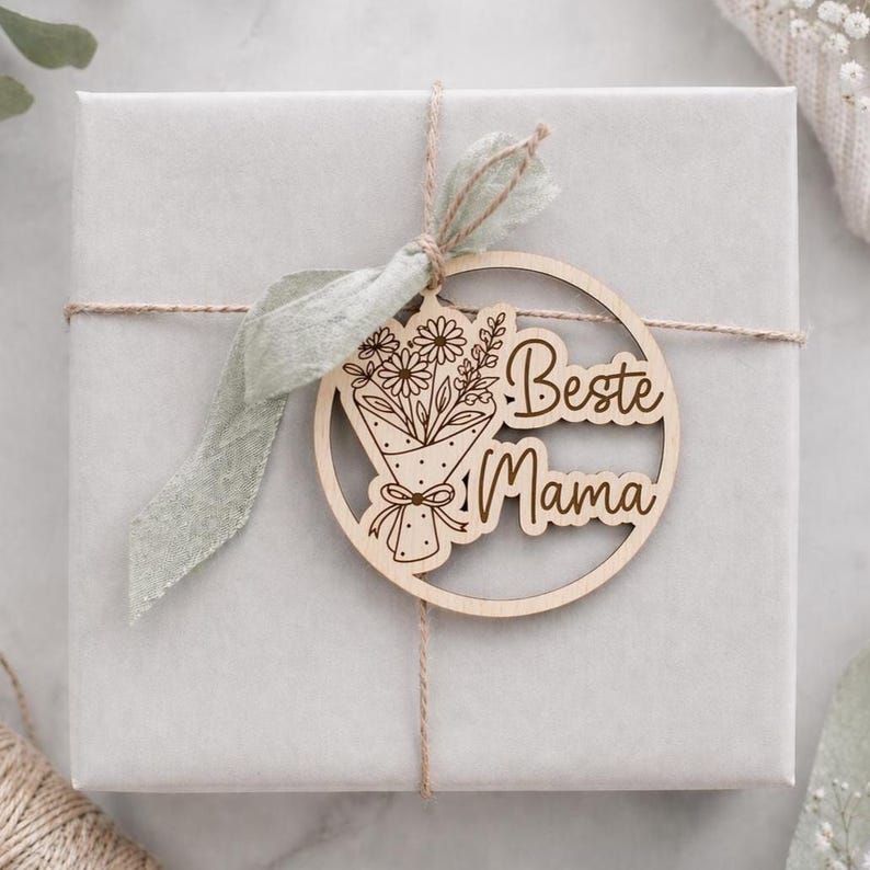 May include: A light gray gift box tied with twine and a sage green ribbon. A wooden circular tag with a laser-cut bouquet of flowers and the words "Beste Mama" is attached to the box. The tag is made of light-colored wood.