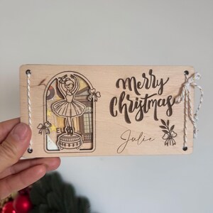 May include: Wooden Christmas card with a ballerina design. The card features the words "Merry Christmas" and the name "Julie" engraved on the front. The card is tied with a white string.