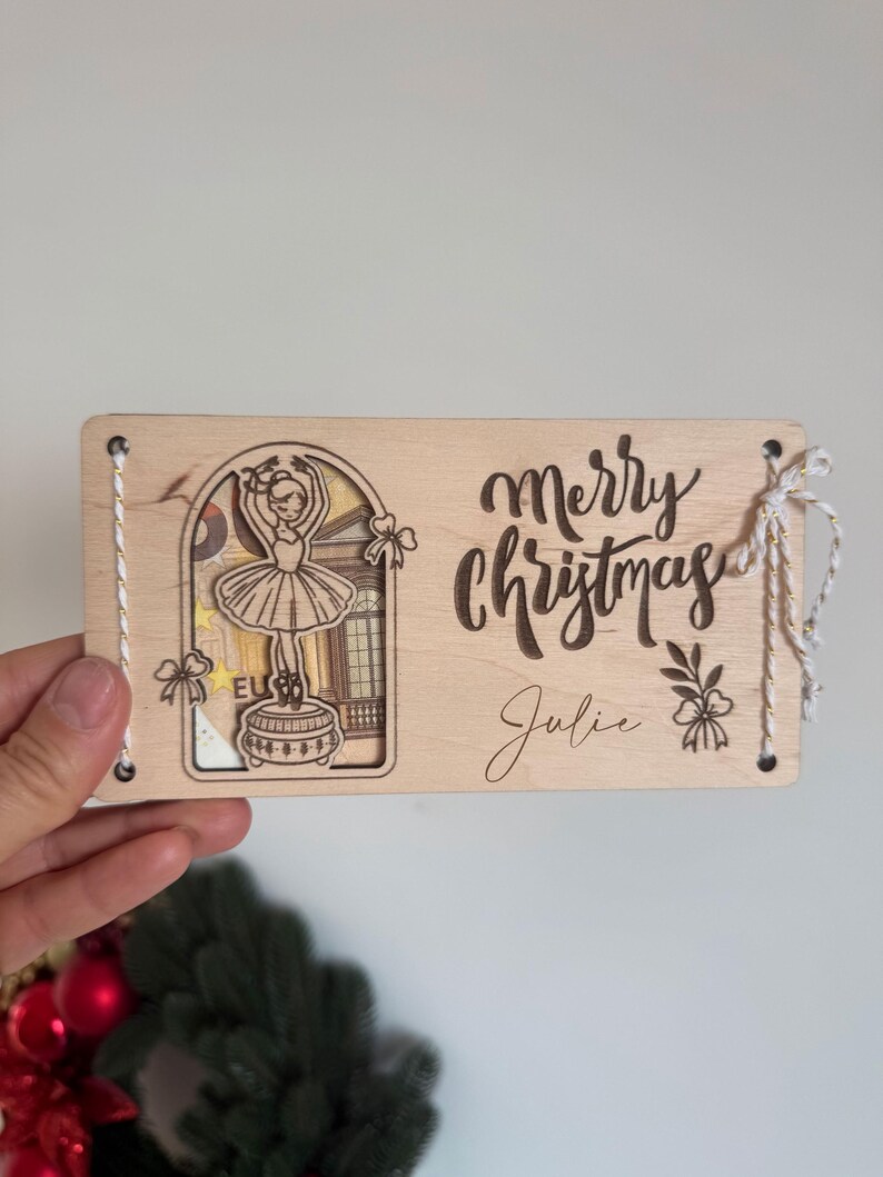 May include: Wooden Christmas card with a ballerina design. The card features the words "Merry Christmas" and the name "Julie" engraved on the front. The card is tied with a white string.