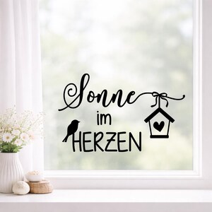 May include: Black vinyl decal with the words "Sonne im Herzen" (Sun in the Heart) in a decorative script. A silhouette of a bird and a birdhouse with a heart are also included in the design. The decal is applied to a window.