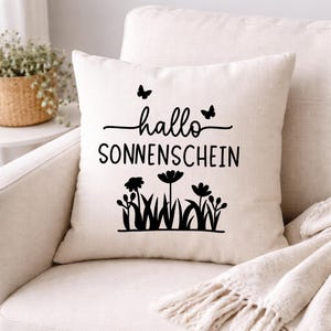 May include: Cream-colored square pillow with the words "hallo SONNENSCHEIN" in black script and a floral design. Two butterflies are above the word "hallo". The pillow is on a cream-colored chair.
