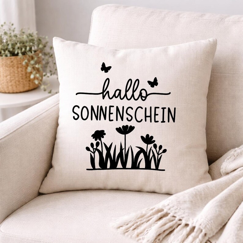 May include: Cream-colored square pillow with the words "hallo SONNENSCHEIN" in black script and a floral design. Two butterflies are above the word "hallo". The pillow is on a cream-colored chair.
