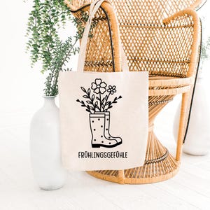 May include: A beige tote bag with a black graphic of flowers and leaves in a polka-dotted rain boot. The text "FR&Uuml;HLINGSGEF&Uuml;HLE" is printed in black below the boot. The bag has two beige handles and is displayed in front of a wicker chair.