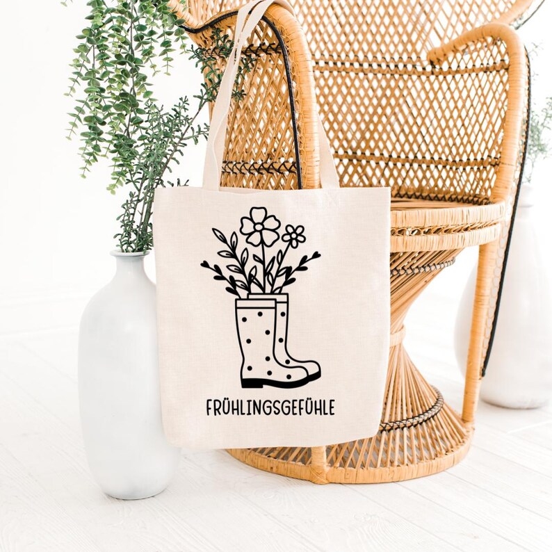 May include: A beige tote bag with a black graphic of flowers and leaves in a polka-dotted rain boot. The text "FR&Uuml;HLINGSGEF&Uuml;HLE" is printed in black below the boot. The bag has two beige handles and is displayed in front of a wicker chair.