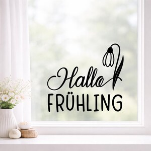 Pode incluir: Autocolante de vinil preto com as palavras "Hallo FR&Uuml;HLING" e um desenho de flor de neve. O autocolante &eacute; aplicado a uma janela, com uma cena exterior desfocada vis&iacute;vel atrav&eacute;s do vidro. O texto est&aacute; numa fonte decorativa.