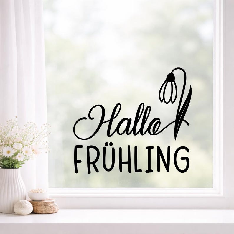 Pode incluir: Autocolante de vinil preto com as palavras "Hallo FR&Uuml;HLING" e um desenho de flor de neve. O autocolante &eacute; aplicado a uma janela, com uma cena exterior desfocada vis&iacute;vel atrav&eacute;s do vidro. O texto est&aacute; numa fonte decorativa.