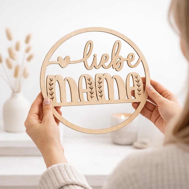 K&ouml;nnte beinhalten: Ein rundes Holzschild mit den Worten "liebe mama" in einer dekorativen Schriftart ausgeschnitten. Das Schild hat Herz- und Bl&auml;tterakzente. Das Schild wird von einer Person gehalten.