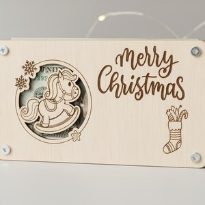 May include: A light-colored wooden money holder with a rocking horse design and the words "Merry Christmas" engraved on the front. The holder is secured with four silver screws and features a slot for inserting money. A Christmas stocking and snowflakes are also engraved.