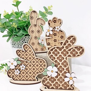May include: Three wooden Easter bunny figurines with floral patterns and white flowers. The bunnies are standing on a wooden base.