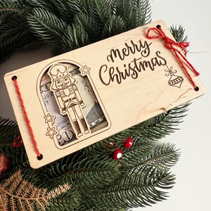 May include: A wooden Christmas gift card holder with a laser-cut nutcracker design and the words "Merry Christmas." The holder is decorated with red string and a red bow, and is set on a bed of evergreen branches.