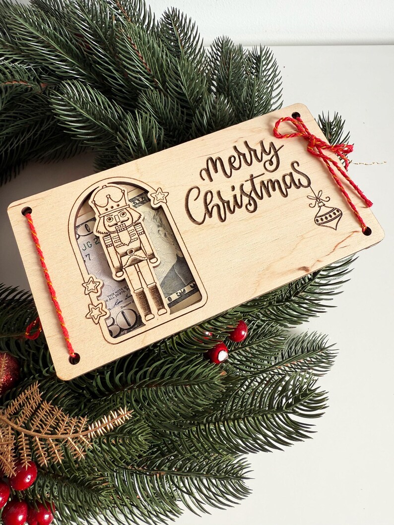 May include: A wooden Christmas gift card holder with a laser-cut nutcracker design and the words "Merry Christmas." The holder is decorated with red string and a red bow, and is set on a bed of evergreen branches.